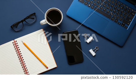 Minimalist flat lay of a blue laptop, open notebook, smartphone, coffee, and office supplies on a dark blue background, symbolizing organized work, study, and technology, perfect for productivity blog 130592120