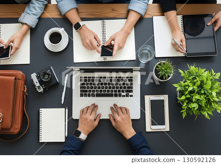 Overhead view of multiple people collaborating on laptops, tablets, and smartphones on a modern dark office desk, ideal for business, technology, and teamwork-related content. 130592126
