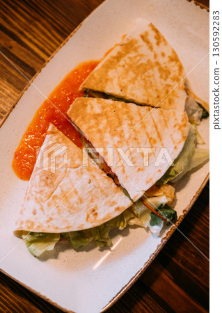 Tortilla food sliced. Served burrito. Grilled stuffed flatbread with lettuce. Hot meal cut piece with red spicy sauce on dish table. 130592283