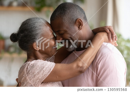 Happy senior couple embracing tenderly indoors, their foreheads touching as they smile with eyes closed, sharing a warm and intimate moment of love and connection 130595568
