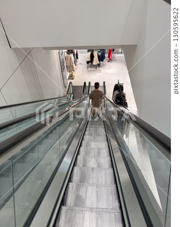 A person descends on a glass escalator inside a modern shopping mall retail space. Consumer movement, urban lifestyle, shopping experience and architectural design within commercial culture. A person descends on a glass escalator inside a modern shopping mall retail space. Consumer movement, urban lifestyle, shopping experience and architectural design within commercial culture. 130595622