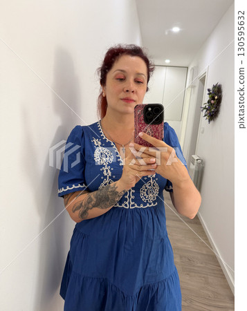 A woman in a blue embroidered dress takes a mirror selfie inside a bright hallway. Identity, self-expression, body image, lifestyle and modern culture reflected through everyday fashion and technology 130595632