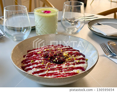 Bowl of creamy oat pudding with raspberry sauce and crushed pistachios served on table. Dessert, sweet food, and culinary creativity in modern dining concept. Bowl of creamy oat pudding with raspberry sauce and crushed pistachios served on table. Dessert, sweet food, and culinary creativity in modern dining concept. 130595654