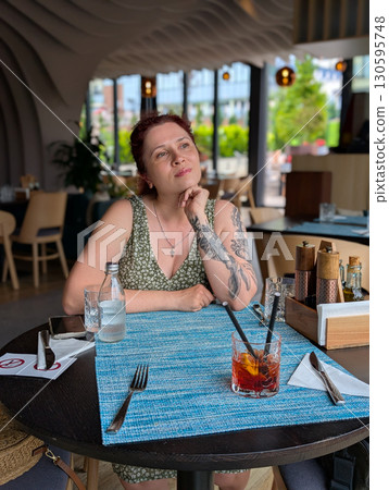 A woman sitting at a restaurant table with a cocktail. Lifestyle, dining, creativity, and relaxed urban atmosphere. A woman sitting at a restaurant table with a cocktail. Lifestyle, dining, creativity, and relaxed urban atmosphere. 130595748