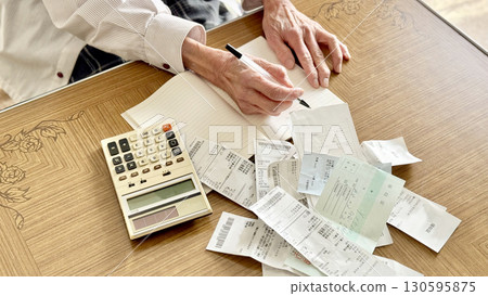 Elderly woman writing in a notebook with a calculator and receipts spread out _ front view 130595875