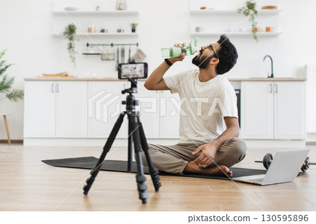 Young man in twenties seated in kitchen recording video about water importance for exercise. Scene includes mat, tripod camera, dumbbell, demonstrating creating fitness content in home environment. 130595896
