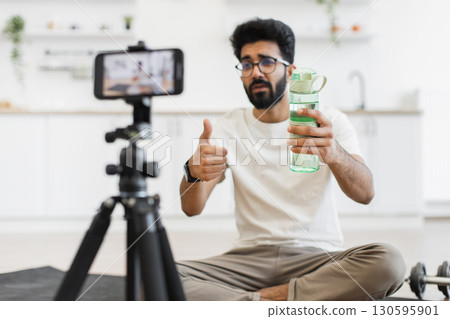 Man in his 30s filming fitness vlog in home kitchen holding water bottle, concept of staying hydrated during workouts, sports, and daily fitness routines, emphasizing health and well-being 130595901