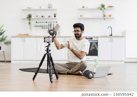 Adult man records fitness video blog at household kitchen, explaining dumbbell exercises. Sitting on floor with camera, tripod, and laptop, demonstrating healthy lifestyle ideas. Adult man records fitness video blog at household kitchen, explaining dumbbell exercises. Sitting on floor with camera, tripod, and laptop, demonstrating healthy lifestyle ideas. 130595942