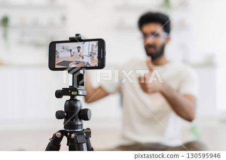 Young man recording video blog using smartphone tripod, demonstrating dumbbell exercises indoors, sharing health tips 130595946