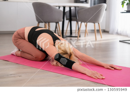 Middle aged blonde woman in sportswear performing stretching exercise on yoga mat at home, improving body flexibility and wellness 130595949