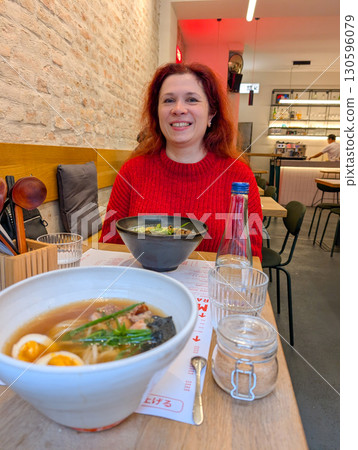 A woman in a red sweater smiles at a table with ramen bowls inside a restaurant. Dining culture, lifestyle, gastronomy and modern culinary tradition in urban hospitality. A woman in a red sweater smiles at a table with ramen bowls inside a restaurant. Dining culture, lifestyle, gastronomy and modern culinary tradition in urban hospitality. 130596079