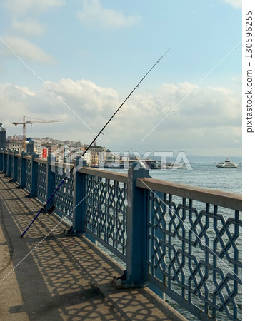 Fishing rod leaning on bridge railing with sea and boats in background. Urban lifestyle, leisure, and traditional activity in city environment. Fishing rod leaning on bridge railing with sea and boats in background. Urban lifestyle, leisure, and traditional activity in city environment. 130596255