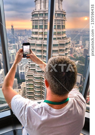 Nov 20, 2019 - Kuala Lumpur, Malaysia: Unrecognizable male adult tourist taking photo of Petronas tower with smartphone through window at colorful sunset.Observation deck visit, tourist attraction Nov 20, 2019 - Kuala Lumpur, Malaysia: Unrecognizable male adult tourist taking photo of Petronas tower with smartphone through window at colorful sunset.Observation deck visit, tourist attraction 130596438