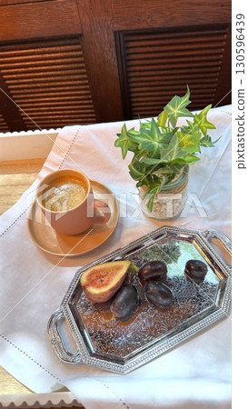 Pastel pink coffee cup resting on saucer beside silver tray with ripe figs, plums, delicate greenery complementing soft morning aesthetic Pastel pink coffee cup resting on saucer beside silver tray with ripe figs, plums, delicate greenery complementing soft morning aesthetic 130596439