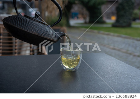 Pouring green tea into a glass from a metal kettle on blurred background, hot drink in teapot 130598924