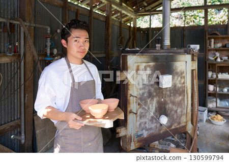 A young potter in an apron standing in front of a kiln A young potter in an apron standing in front of a kiln 130599794