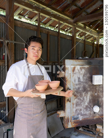 A young potter in an apron standing in front of a kiln A young potter in an apron standing in front of a kiln 130599796