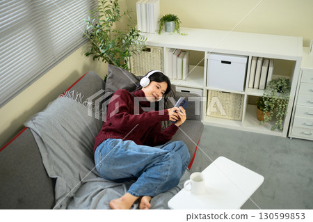 Young woman relaxing on a sofa at home, wearing headphones and using smartphone Young woman relaxing on a sofa at home, wearing headphones and using smartphone 130599853