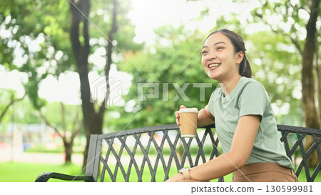 Smiling young woman relaxing on a park bench while holding a takeaway coffee cup 130599981