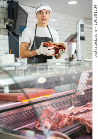 Positive young salesman demonstrating piece of meat in butcher shop Positive young salesman demonstrating piece of meat in butcher shop 130601234