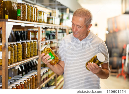 Mature man in supermarket selects examines compares two cans of olives. 130601254