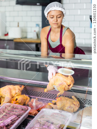 In butcher shop, young female salesperson in gloves shows damp dressed chicken 130601330