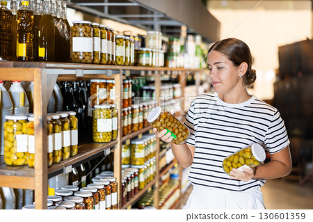 Young woman in supermarket selects examines compares two cans of olives. 130601359