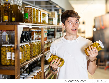Interested guy choosing pickled olives in glass jars in supermarket 130601432