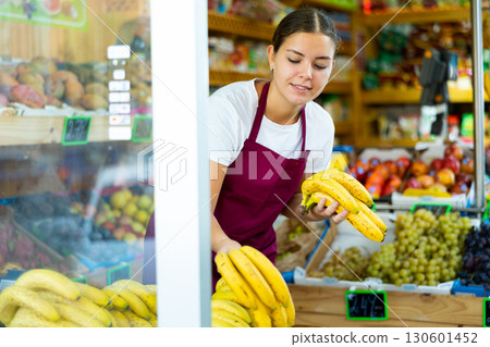Positive young European woman in apron picking out ripe bananas from wooden box in hypermarket Positive young European woman in apron picking out ripe bananas from wooden box in hypermarket 130601452