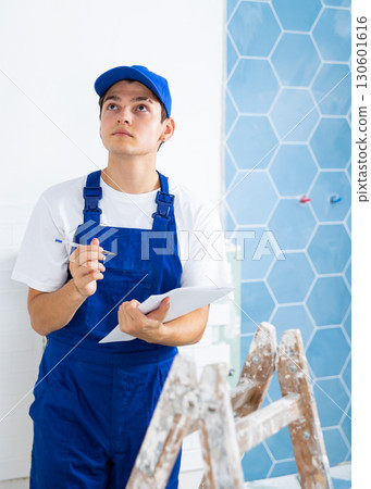 Portrait of man builder in construction overalls with documents in hands checks quality of completed repairs in apartment Portrait of man builder in construction overalls with documents in hands checks quality of completed repairs in apartment 130601616