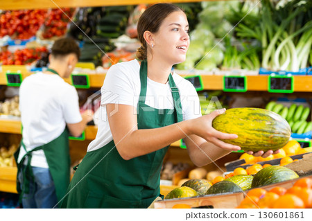 Cheerful young female supermarket worker in apron holding out ripe melon in fruit section Cheerful young female supermarket worker in apron holding out ripe melon in fruit section 130601861