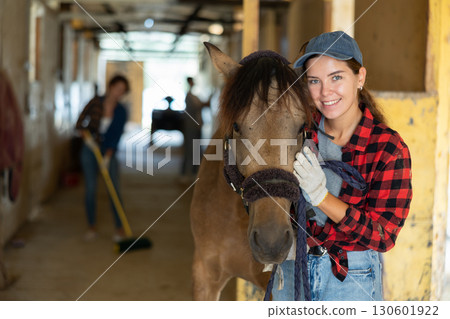 Female owner of horse takes animal out of stall, holds bridle and forces it to follow her 130601922