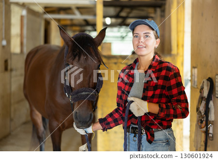 Female owner of horse takes animal out of stall, holds bridle and forces it to follow her 130601924