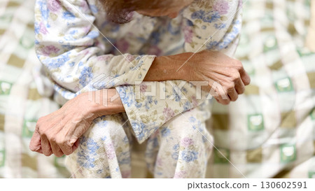An elderly woman wearing pajamas and sitting on a bed with her arms folded facing downwards 130602591