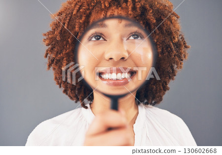 Funny, smile and black woman with a magnifying glass on face isolated on a grey studio background. Happy, comic and facial zoom of a girl with a magnifier for comedy, happiness and discovery 130602668