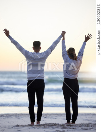 Couple, beach and hands up outdoor while happy at sunset for love, freedom and peace with calm ocean. Young man and woman together on vacation holiday at sea to relax, travel and connect in nature 130602858