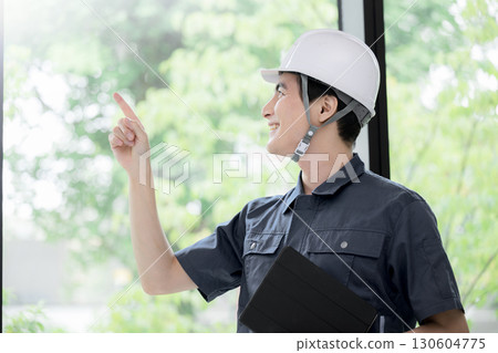 一名身穿工作服、戴著安全帽的男子在綠樹成蔭的窗邊檢查建築物的安全狀況和其他維修工作。 130604775