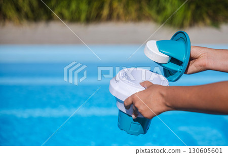 Hands placing chlorine tablet into floating dispenser above clean blue pool water for safe swimming and proper maintenance 130605601