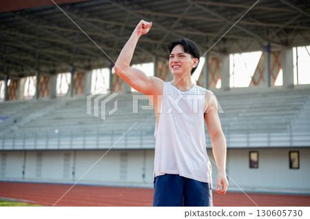 An asian man in white tank top is smiling and raising a fist while standing on racetrack. An asian man in white tank top is smiling and raising a fist while standing on racetrack. 130605730