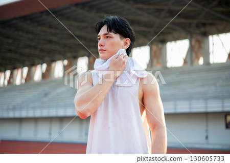 Asian man in white tank top standing and wiping his sweat, taking a rest after running on racetrack. 130605733