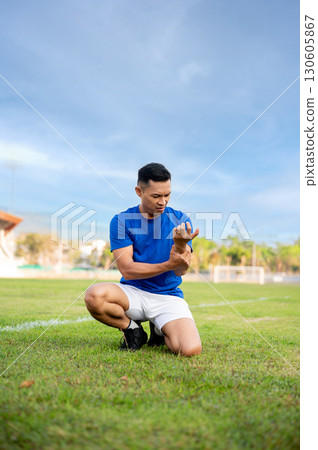 Asian man in blue shirt holding and looking at his arm while kneeling on grass. Arm pain or injury. Asian man in blue shirt holding and looking at his arm while kneeling on grass. Arm pain or injury. 130605867