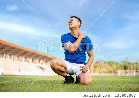 Asian man in blue shirt is kneeling on grass and touching or massaging his shoulder. Pain or injury. Asian man in blue shirt is kneeling on grass and touching or massaging his shoulder. Pain or injury. 130605868