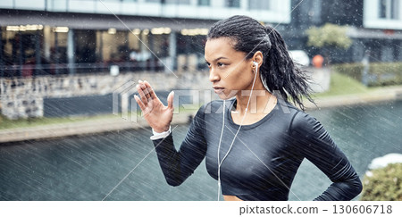 Black woman, fitness and running with earphones in the rain for sports motivation or determination in the city. African American sporty female runner doing intense cardio workout in the rainy weather 130606718