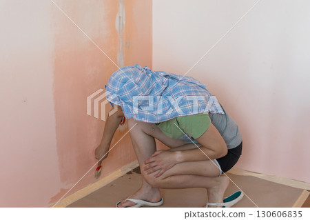 Home Renovation Concept: Woman Painting Peach-Colored Wall in Room Corner with Blue Shirt and Painters Tape. 130606835