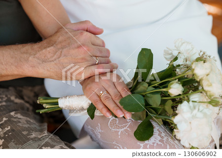 Hands Holding Wedding Rings with White Bouquet, Rustic Detail, Marriage Commitment, Golden Bands, Lace Texture, Warm Colors, Close-Up View. 130606902