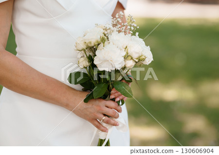 Bridal Bouquet of White Roses Held by Bride in Wedding Dress, Lush Green Field Backdrop, Classic Romantic Celebration. 130606904