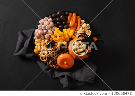 Halloween sweet charcuterie board with pumpkin, popcorn, color chocolate candy, spider, crackers on black background. View from above. Halloween sweet charcuterie board with pumpkin, popcorn, color chocolate candy, spider, crackers on black background. View from above. 130608478