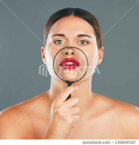 Beauty, magnifying glass and portrait of a woman in studio with a makeup, skincare and natural routine. Face, cosmetic and female model with glass lens on her mouth while isolated by gray background. Beauty, magnifying glass and portrait of a woman in studio with a makeup, skincare and natural routine. Face, cosmetic and female model with glass lens on her mouth while isolated by gray background. 130608587