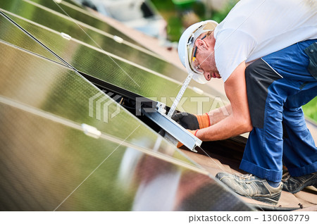 Worker building photovoltaic solar panel system on rooftop of house. Man using ruler to measure mounting equipment for precise installation for generating electricity through photovoltaic effect. 130608779