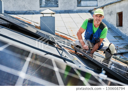 Worker building solar panel system on metal rooftop of house. Portrait of engineer installing photovoltaic solar module outdoors. Alternative, green and renewable energy generation concept. Worker building solar panel system on metal rooftop of house. Portrait of engineer installing photovoltaic solar module outdoors. Alternative, green and renewable energy generation concept. 130608794